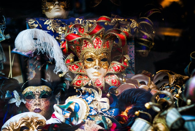 Das Bild zeigt einen Theaterkoffer mit harmonisch bunten venezianischen Masken. Eine in gold-rot bemalte Maske sticht besonders hervor.