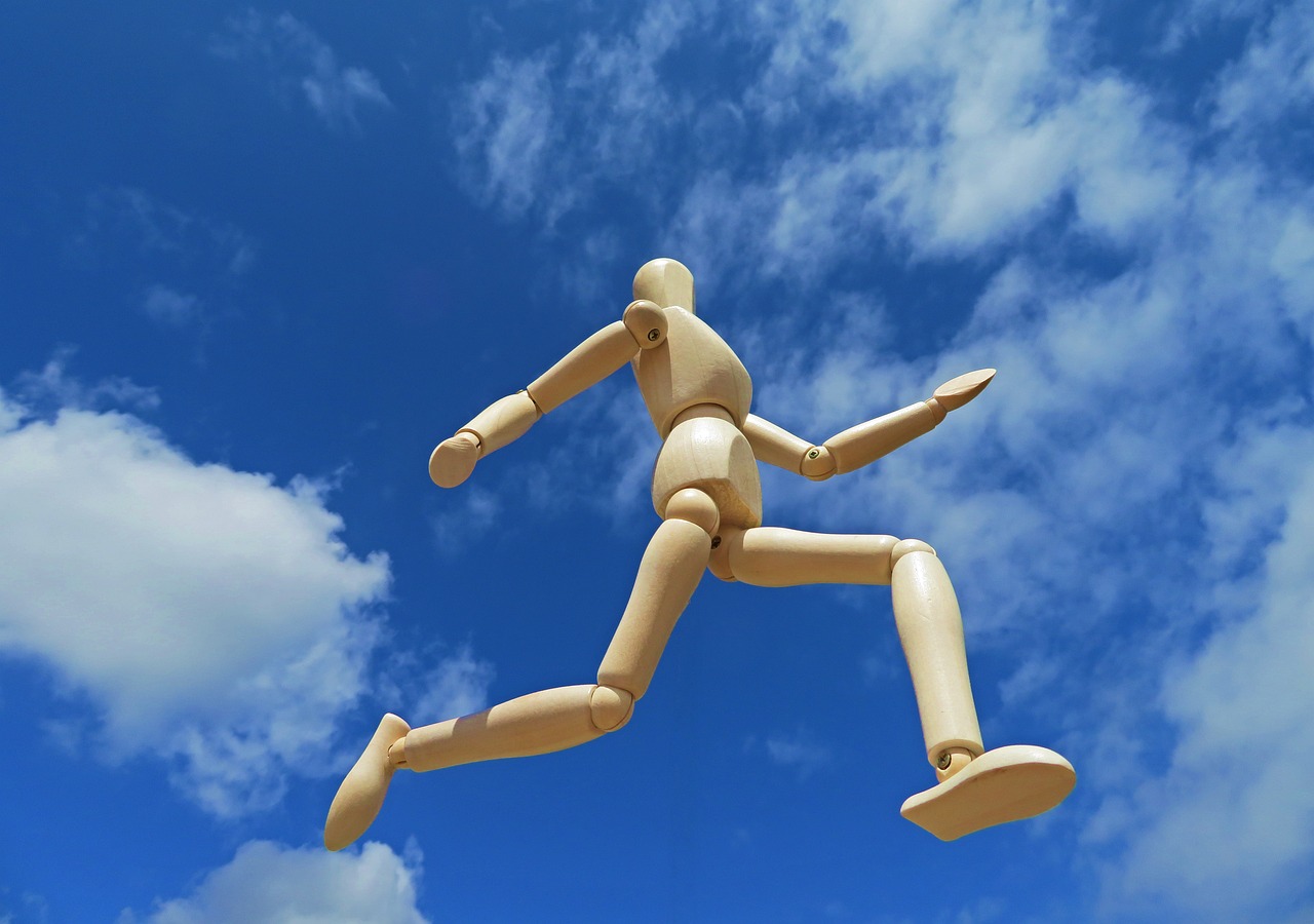 Eine Holzfigur springt schwungvoll dynamisch vor blauem Himmel zurück ins Büro.