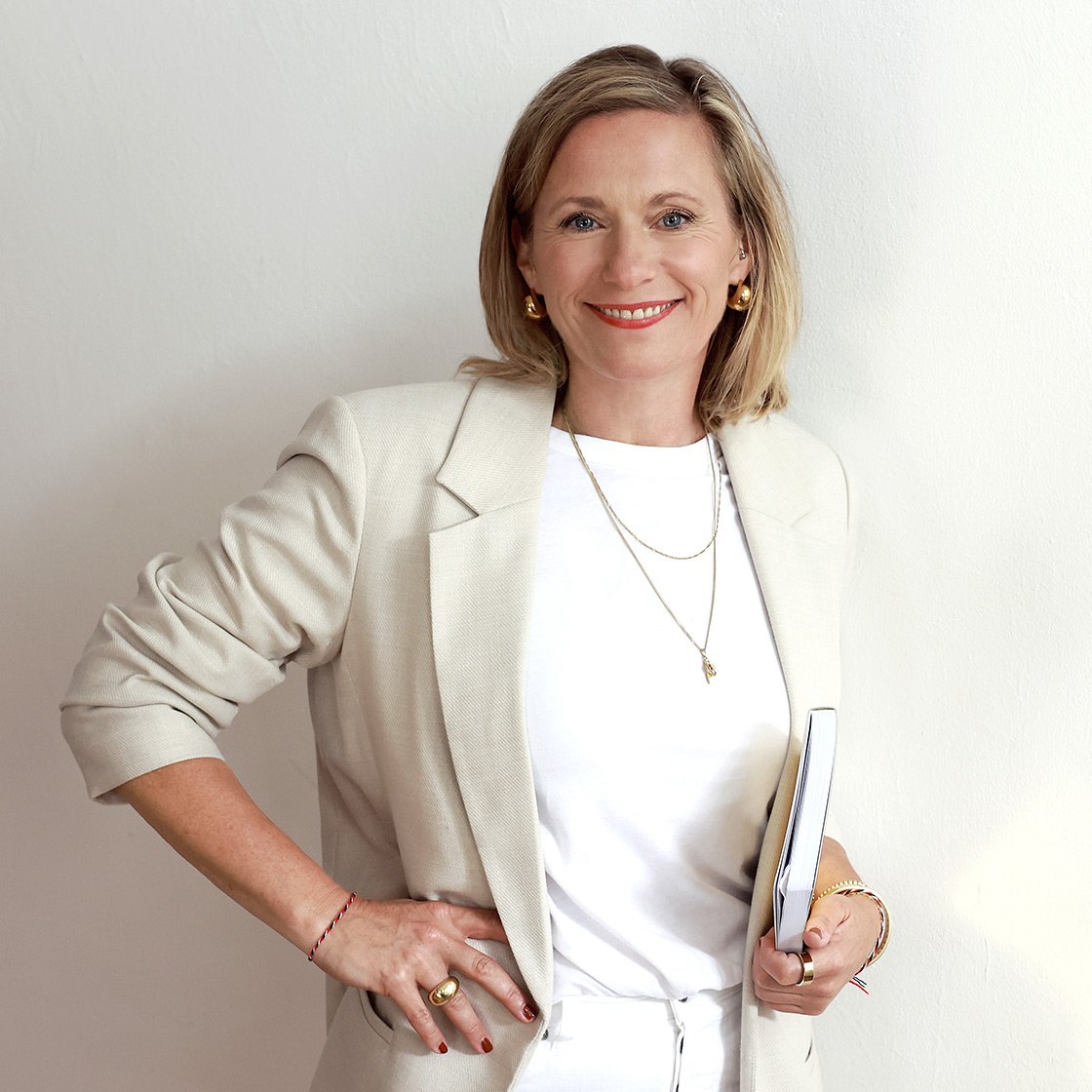 futures-literacy_charlotte_heidsiek_final Charlotte stehend mit Buch unterm Arm vor weißer Wand und in hellem beigefarbenem Jacket.